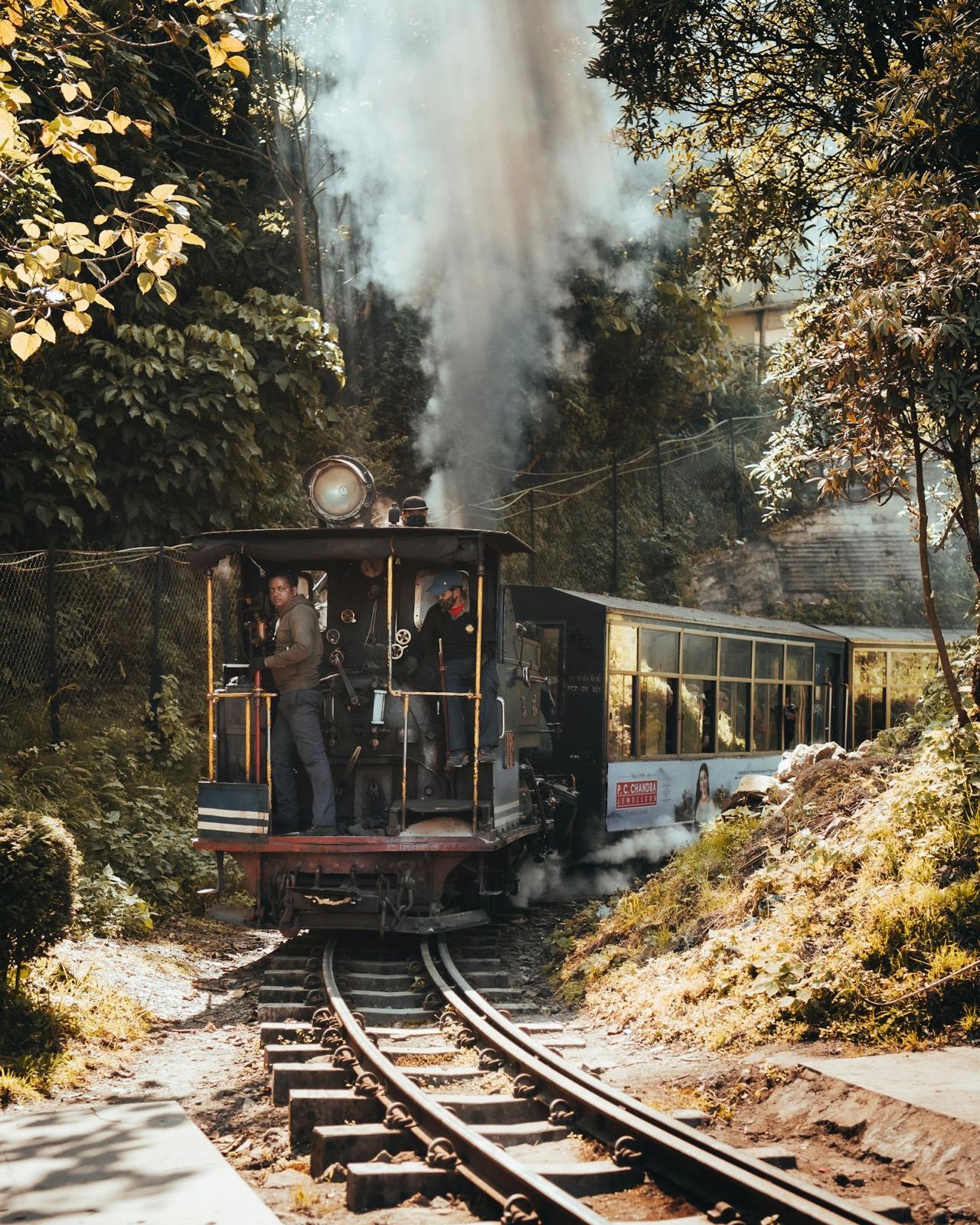 A small steam locomotive with its crew in the cab, pulling a carriage along narrow-gauge track through trees, steam rising behind it