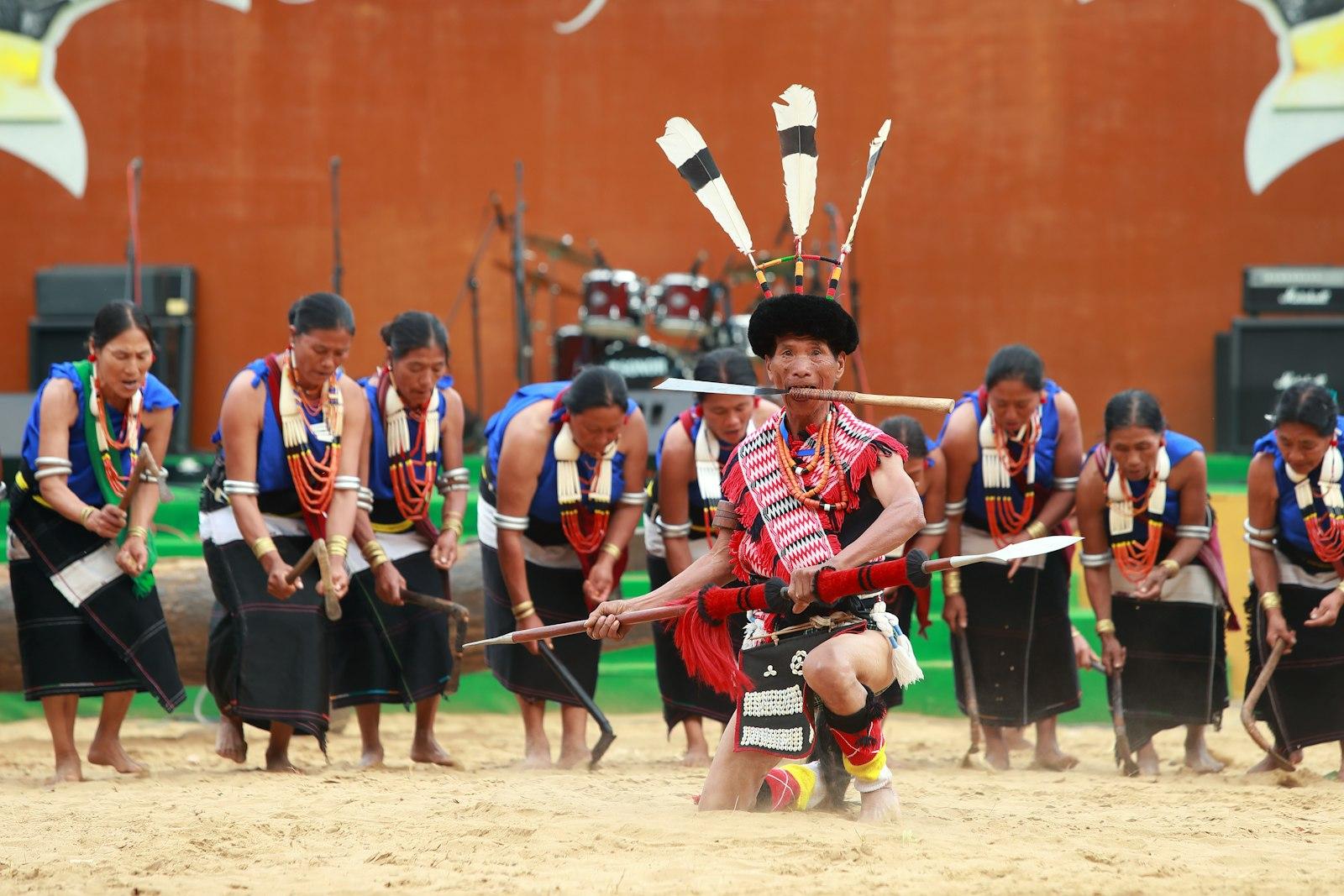 A Naga man in a feathered headdress kneels with a spear and a blade, while a line of women in blue and black dance behind him