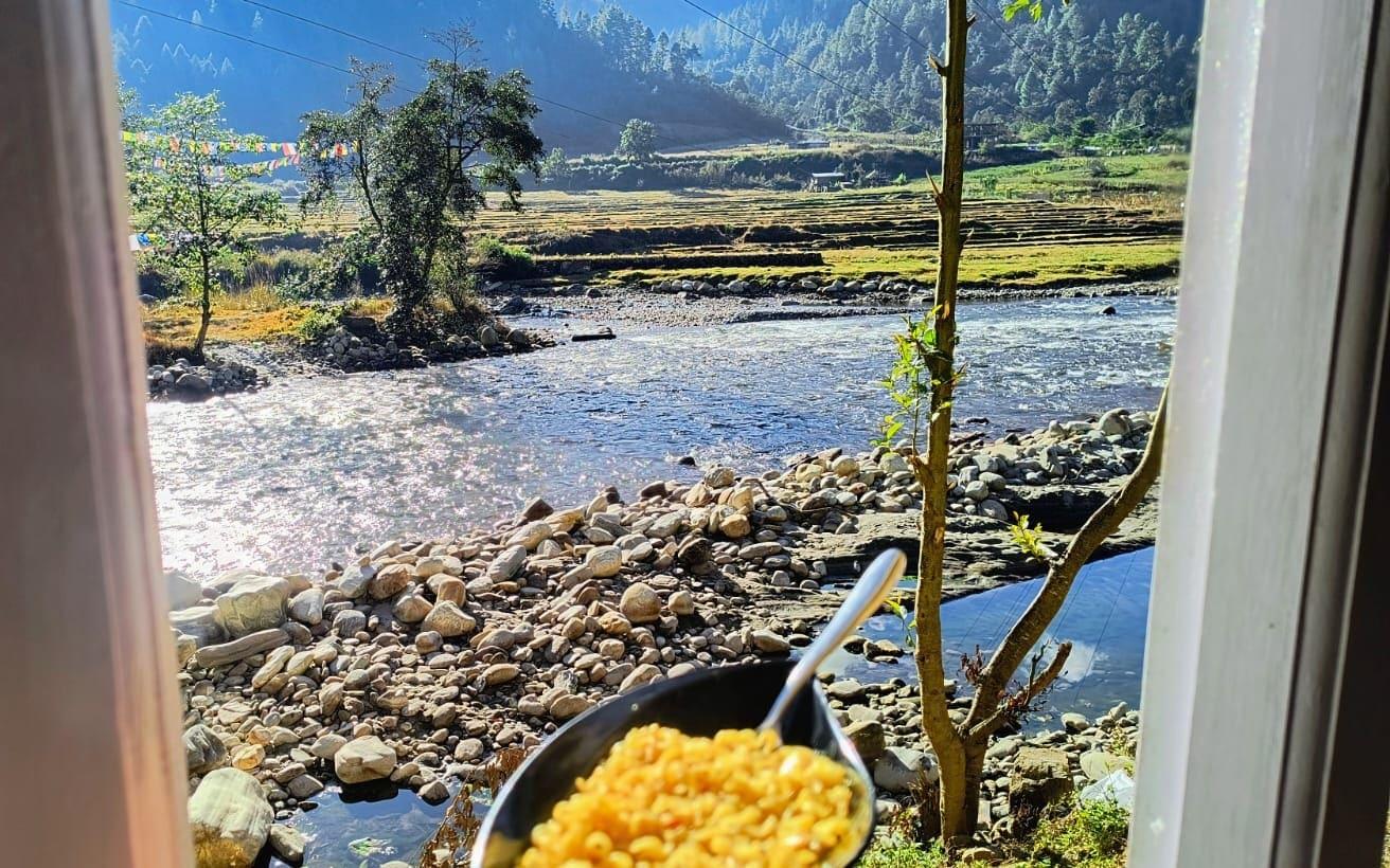 A bowl of breakfast held at a window overlooking a river, terraced fields and pine hills in Arunachal Pradesh